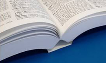 A softcover book lying open on a flat surface, demonstrating the Otabind-style layflat binding where the cover spine stays slightly lifted and detached from the inner pages to prevent closing.